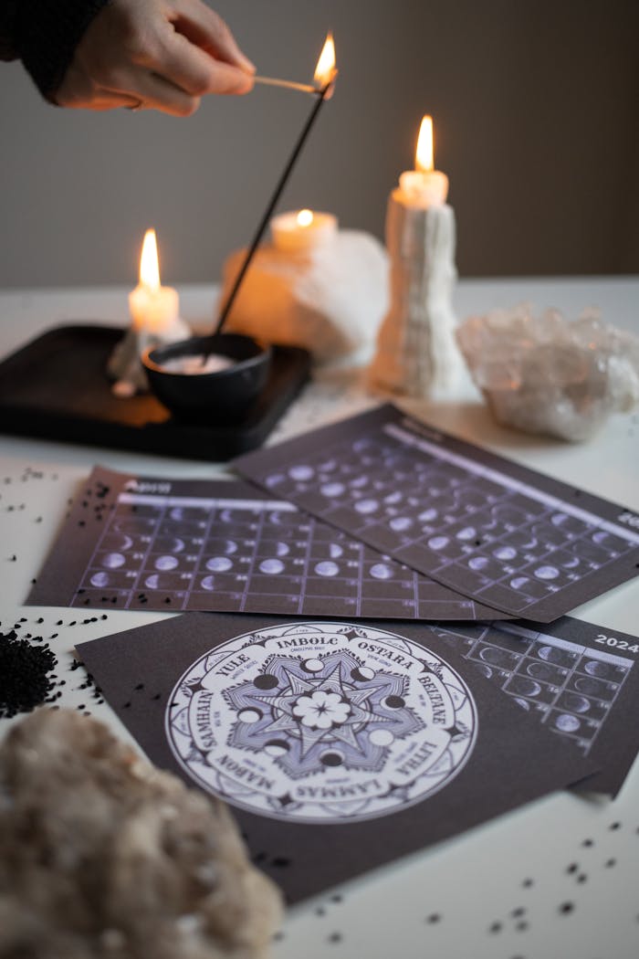 Mystical scene with a hand lighting an incense stick, lit candles, and a moon phase calendar.